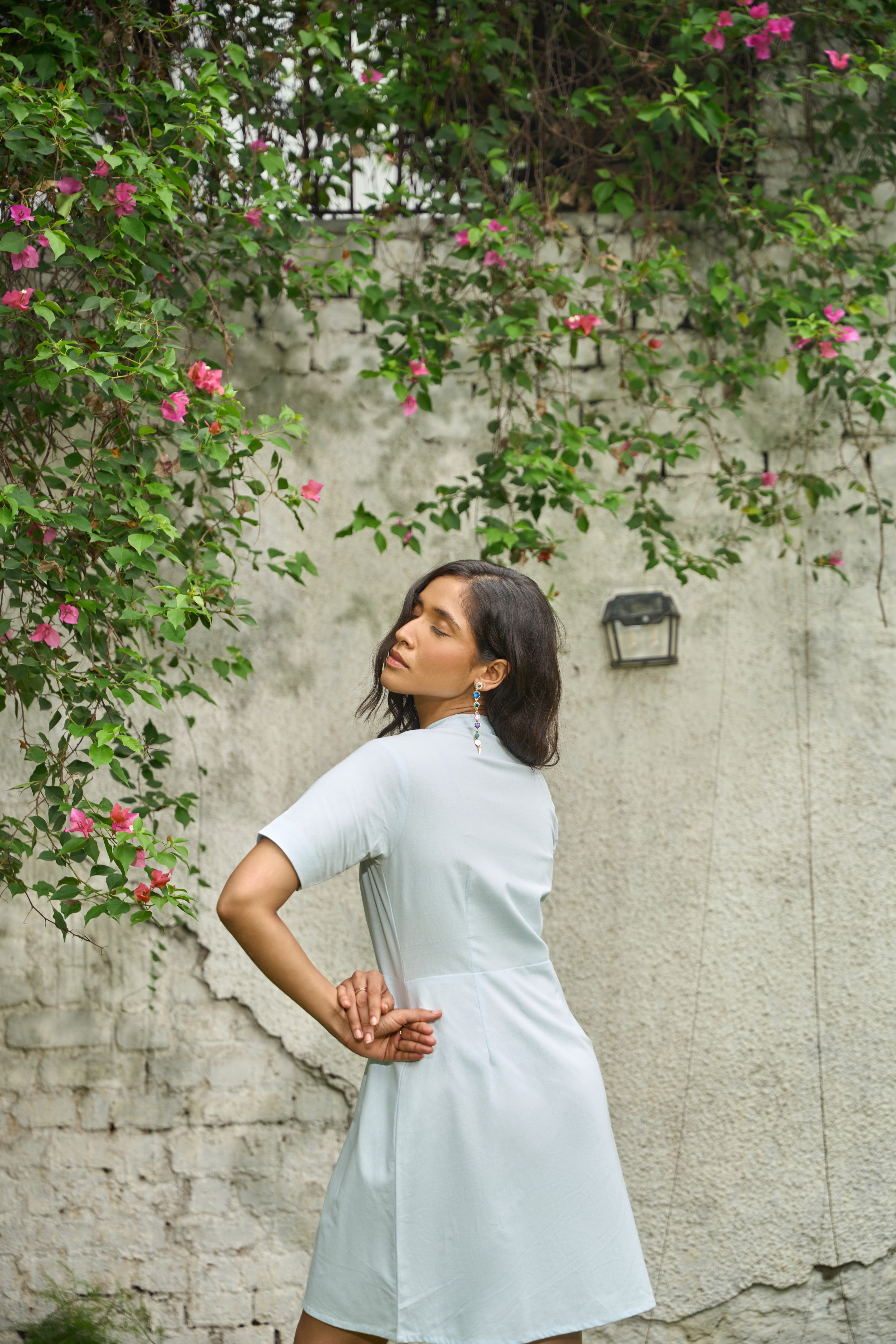 Woman in a light dress standing against a textured wall with greenery and flowers.