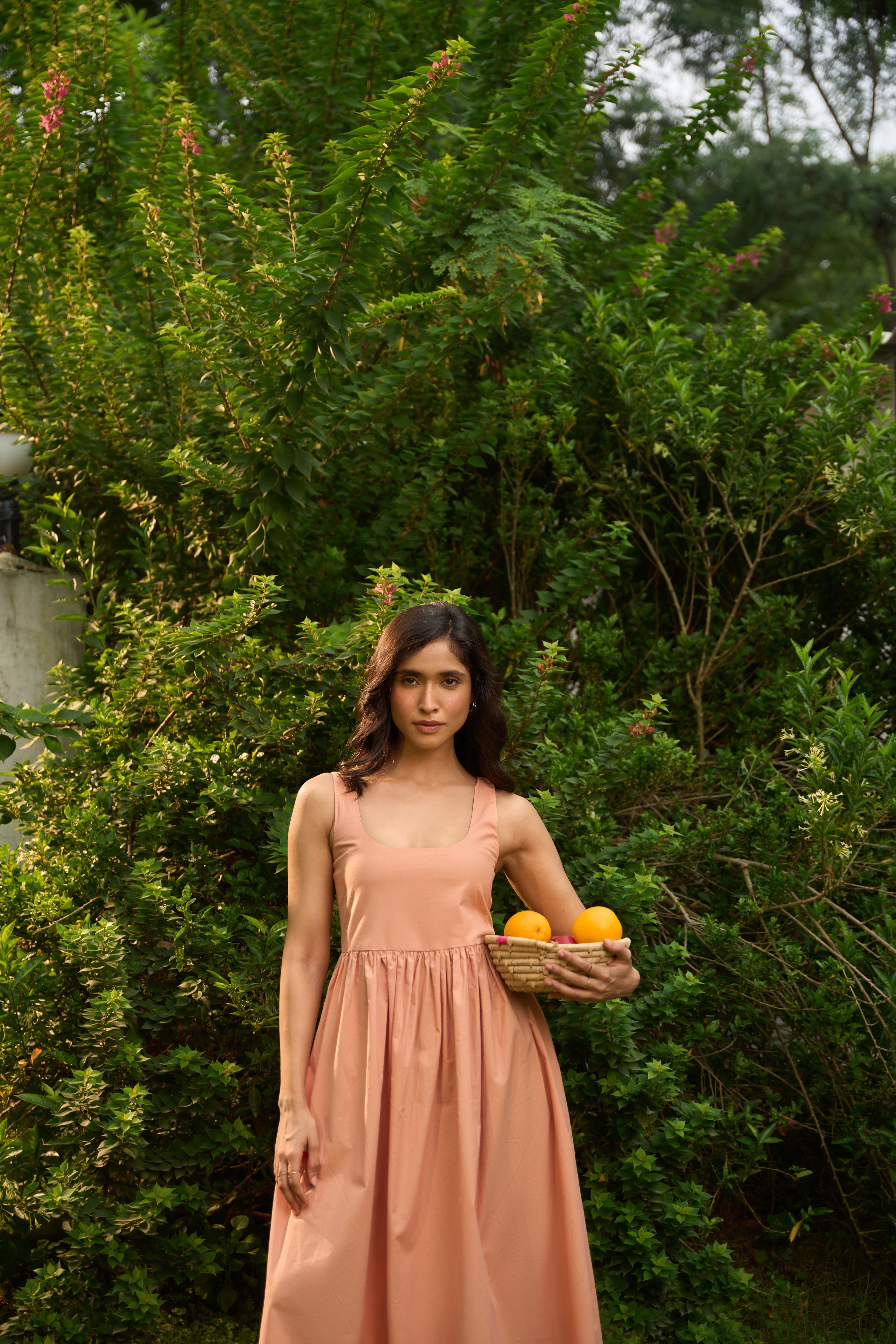Woman in a peach dress holding a basket of oranges in a green outdoor setting